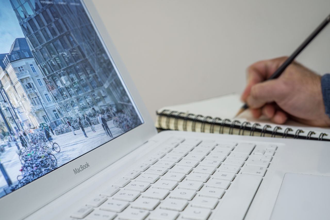 our-story A person taking notes in a notebook beside an open laptop displaying an urban scene.